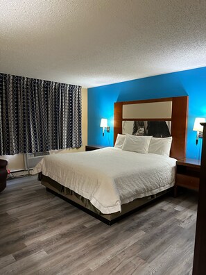 Een hotelkamer met een groot bed, een nachtkastje met een lamp, een stoel en een wandairconditioner.