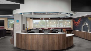 A modern hotel reception area with a curved wooden reception desk, a large window offering a view of the outside, and a wall with a colorful dot pattern. - Tru By Hilton Columbus East Broad (Columbus)
