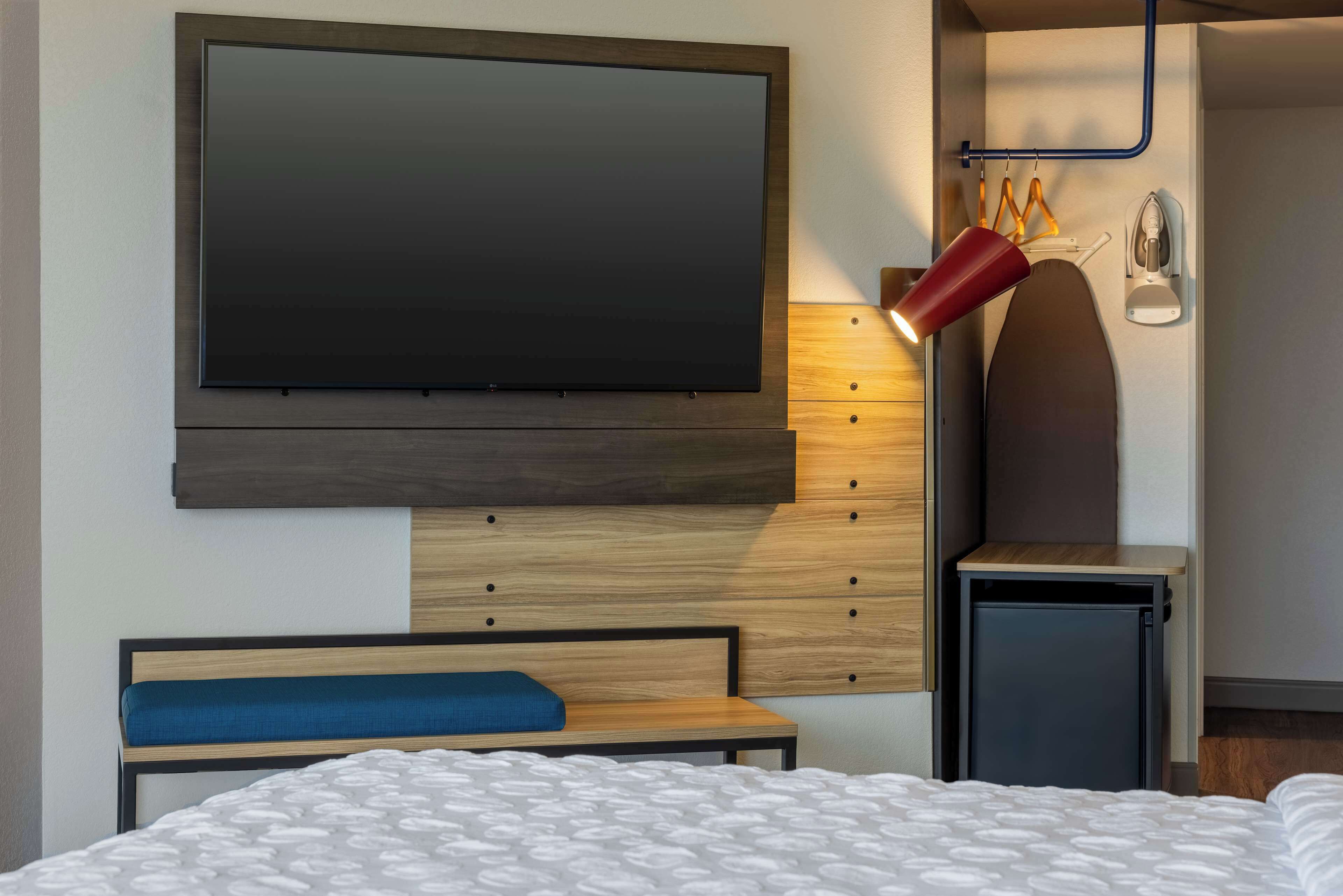 A modern hotel room with a large flat-screen TV mounted on the wall, a bedside table with a lamp, a small desk, and a bed with a white patterned bedspread.