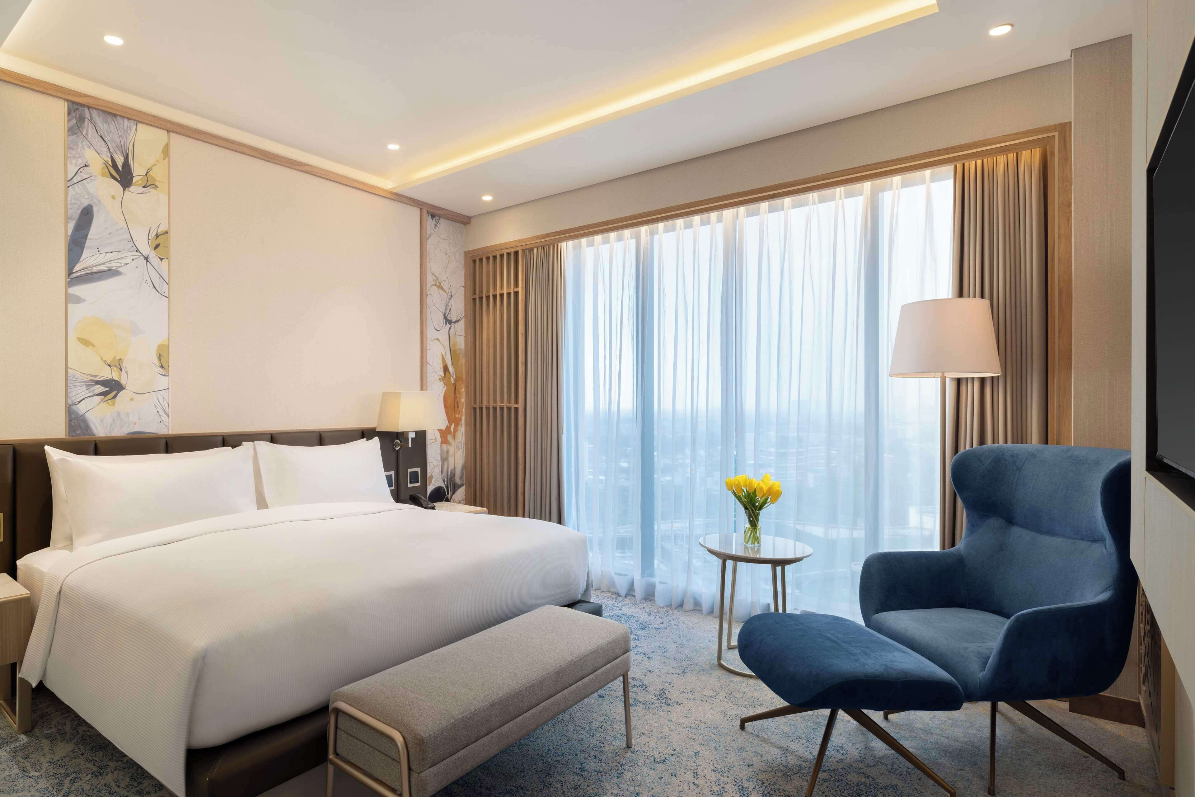A hotel room with a large bed, a bedside table with a lamp, a chair, a small table with a vase of flowers, and a large window with curtains.