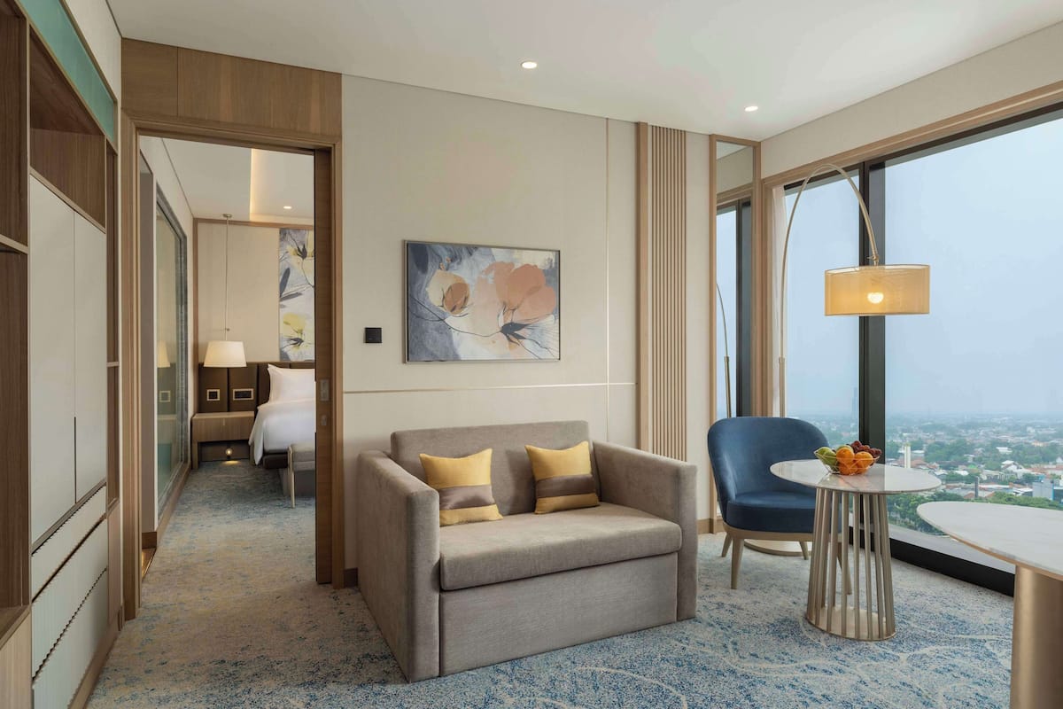 A hotel room with a sofa, a chair, a small table with fruit, and a large window offering a city view.
