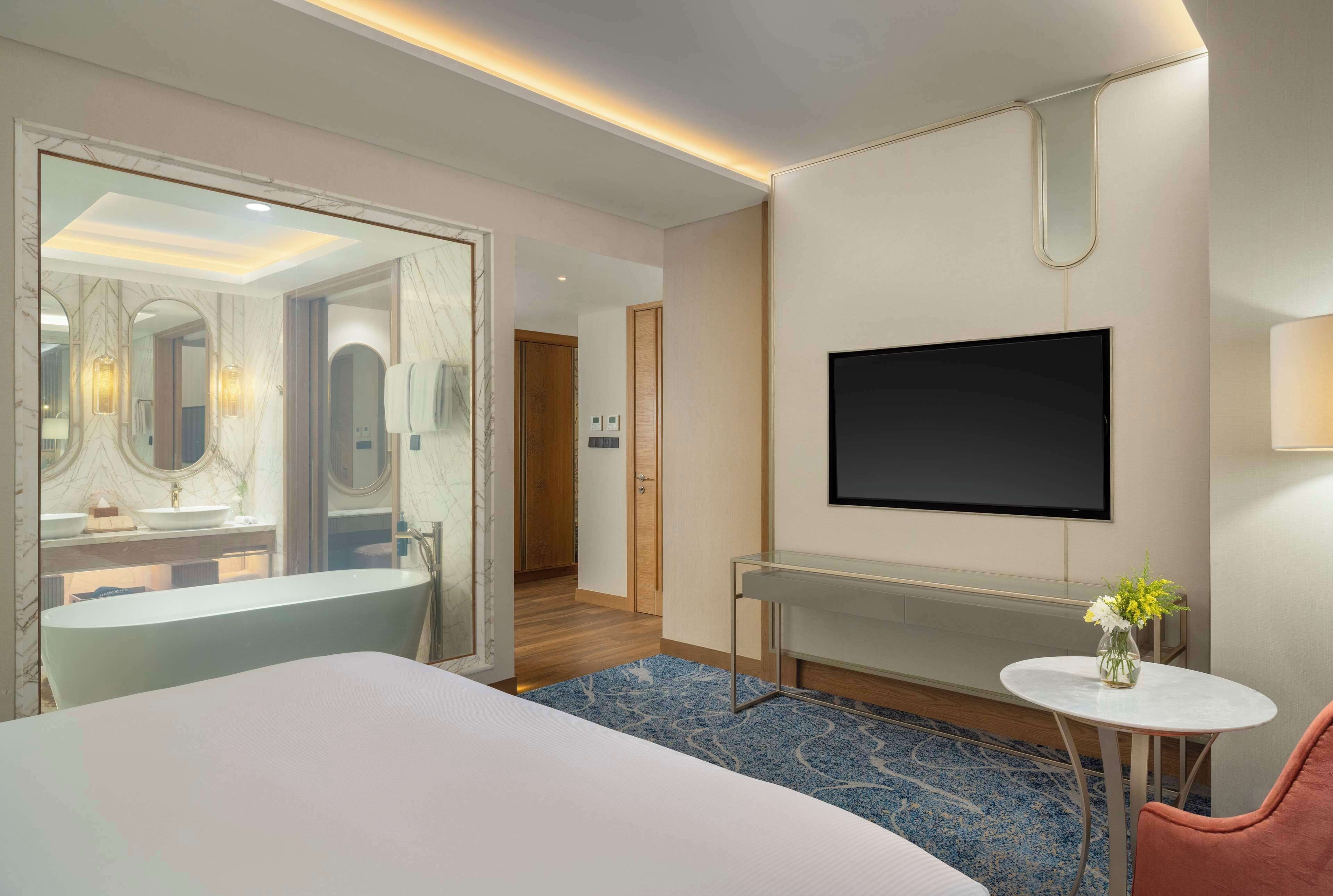 Une chambre d’hôtel moderne avec un grand lit, un téléviseur fixé au mur, une petite table avec un vase et une salle de bain visible à travers une porte ouverte.