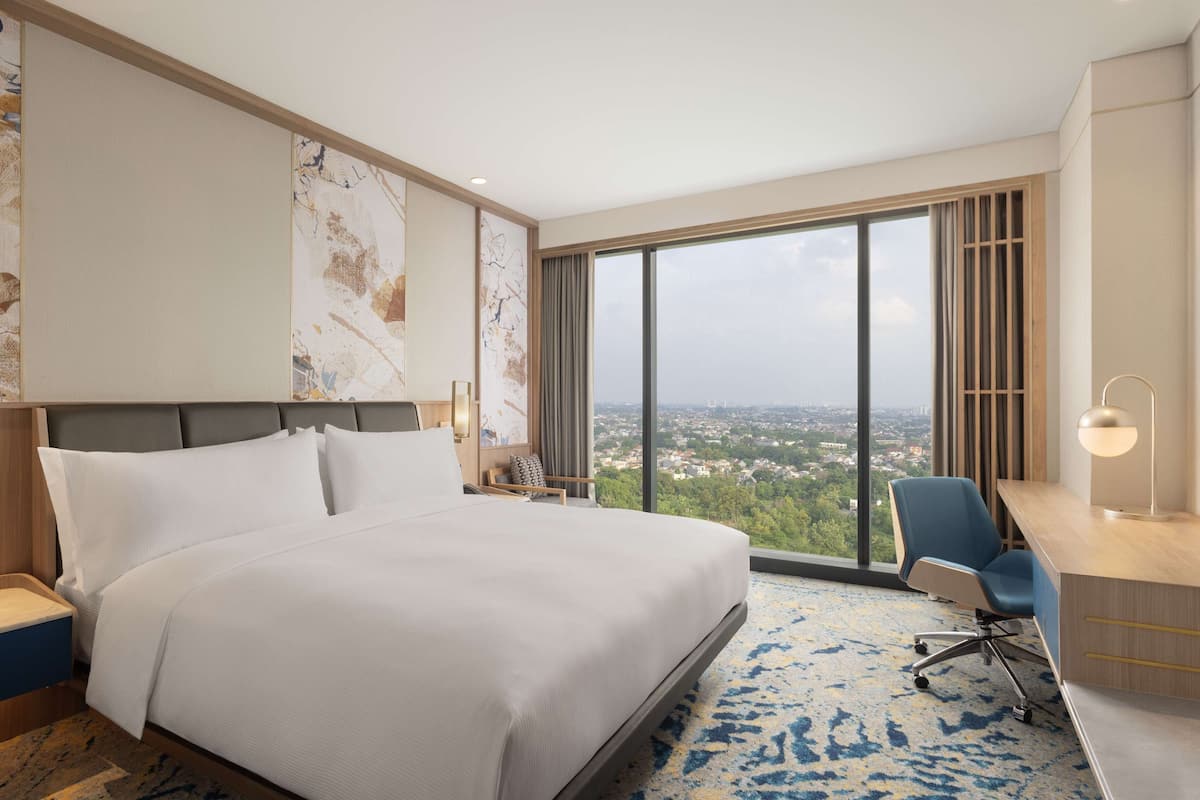 A hotel room with a large bed, a desk with a chair, a TV, and a view of a cityscape through large windows.