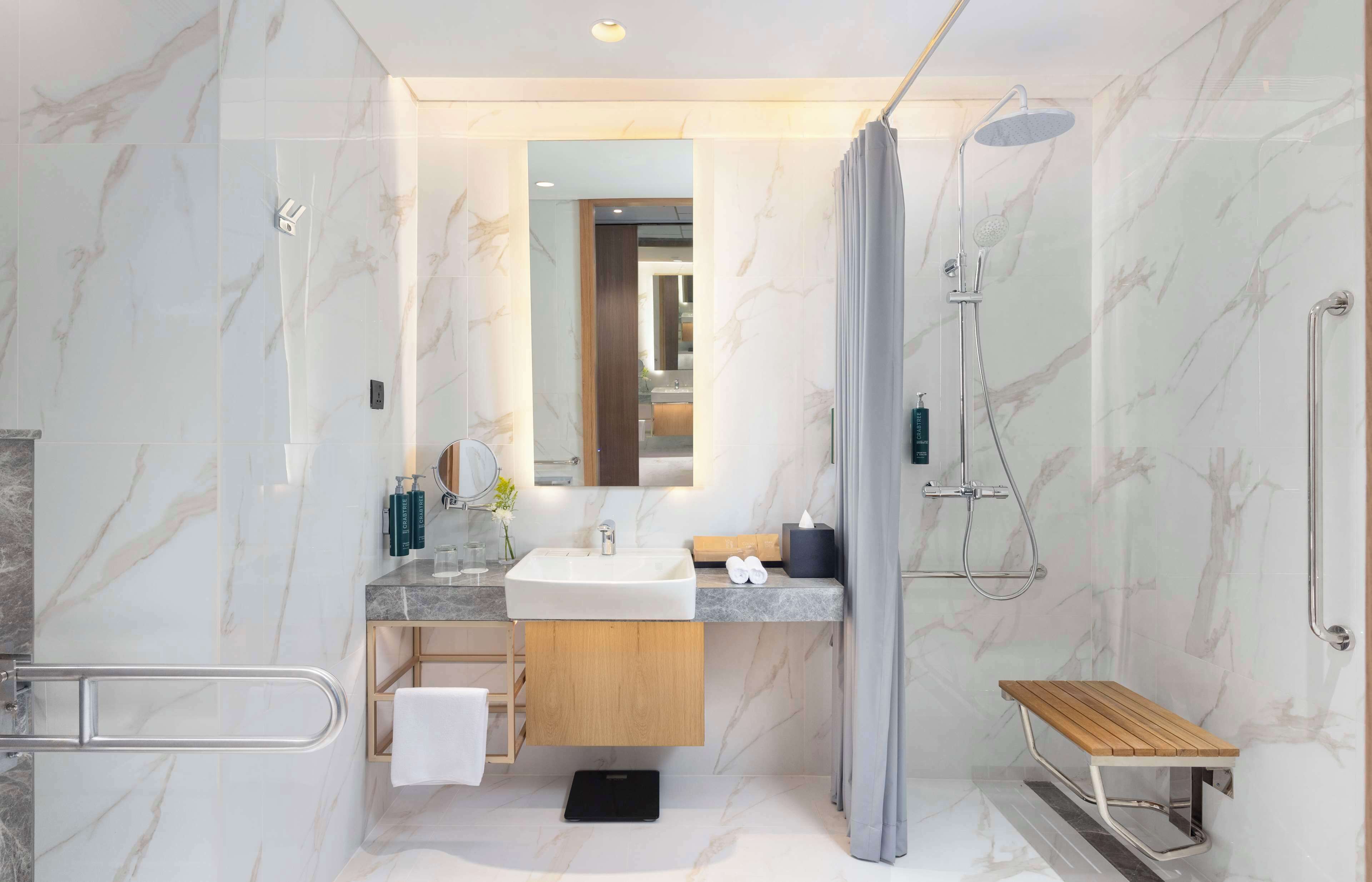 A modern bathroom with a marble wall, a sink, a mirror, a shower, and a wooden bench.