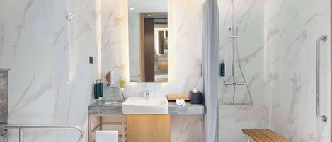 A modern bathroom with a marble wall, a sink, a mirror, a shower, and a wooden bench.