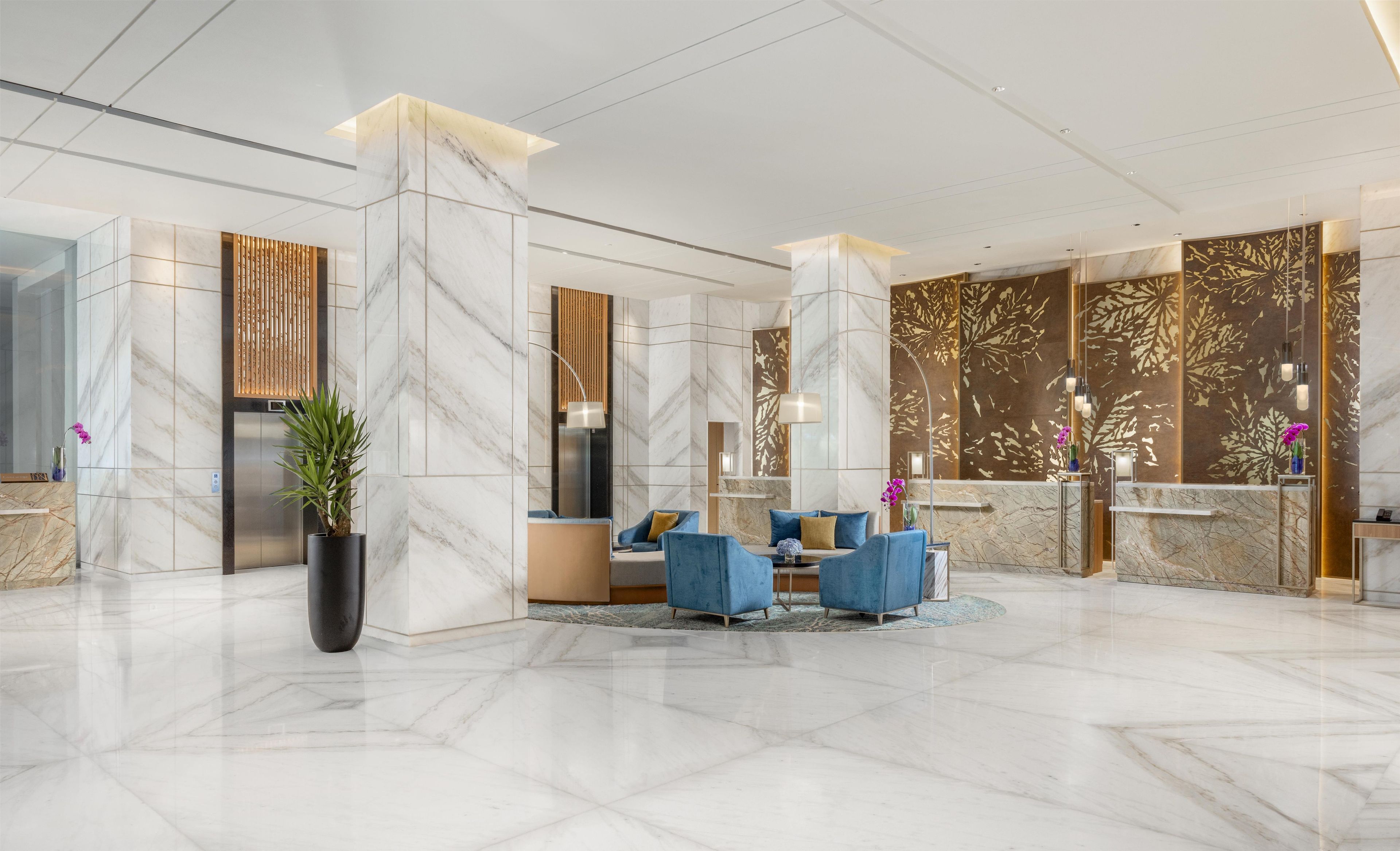 A modern lobby with marble flooring, a reception desk, seating area with blue chairs, and decorative wall panels.