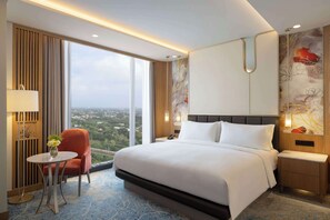 Habitación de hotel con una cama grande, una mesita con un florero, una silla, una mesita de noche y vistas a la ciudad.