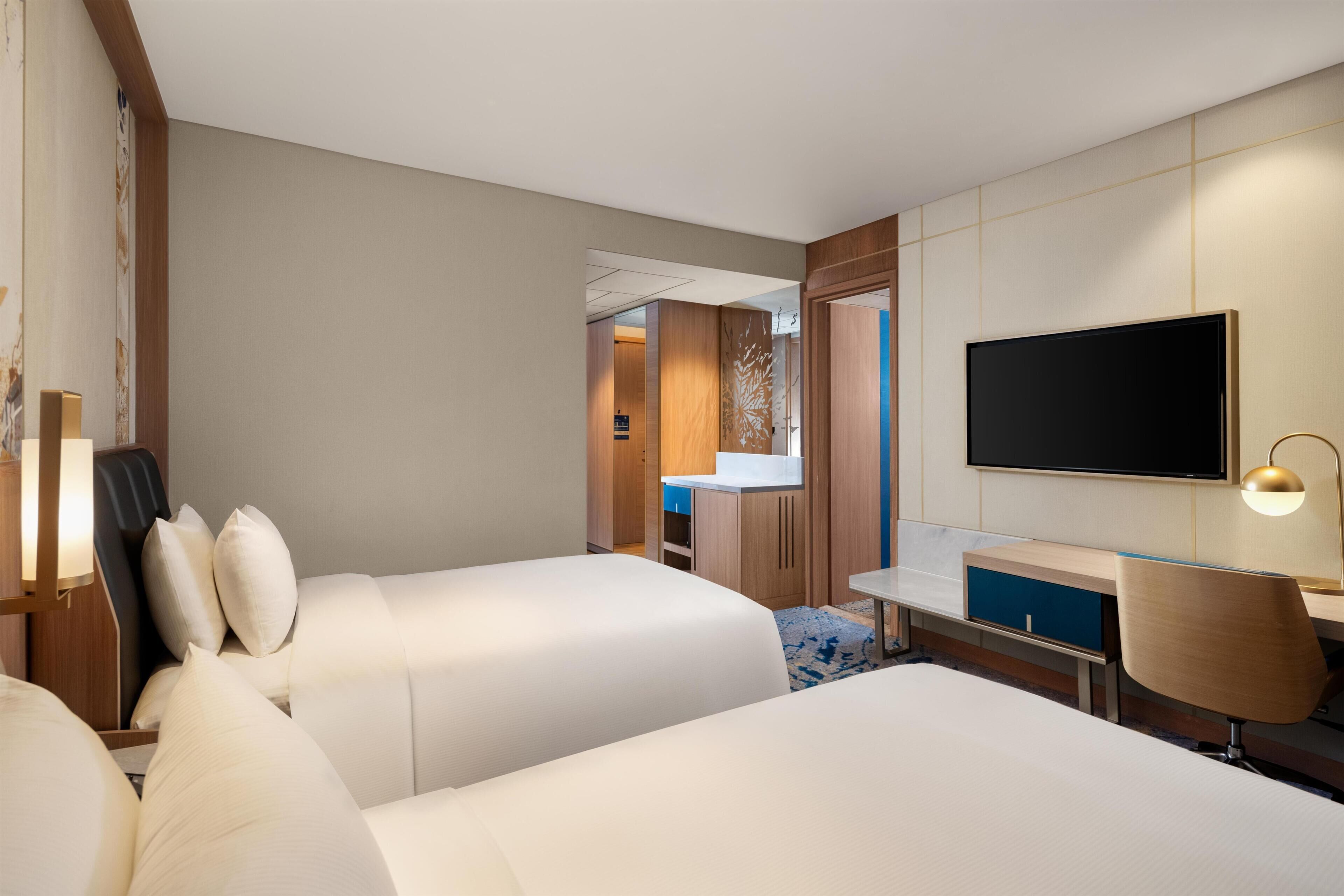 Habitación de hotel con dos camas, un escritorio, una televisión y un baño visible a través de una puerta abierta.