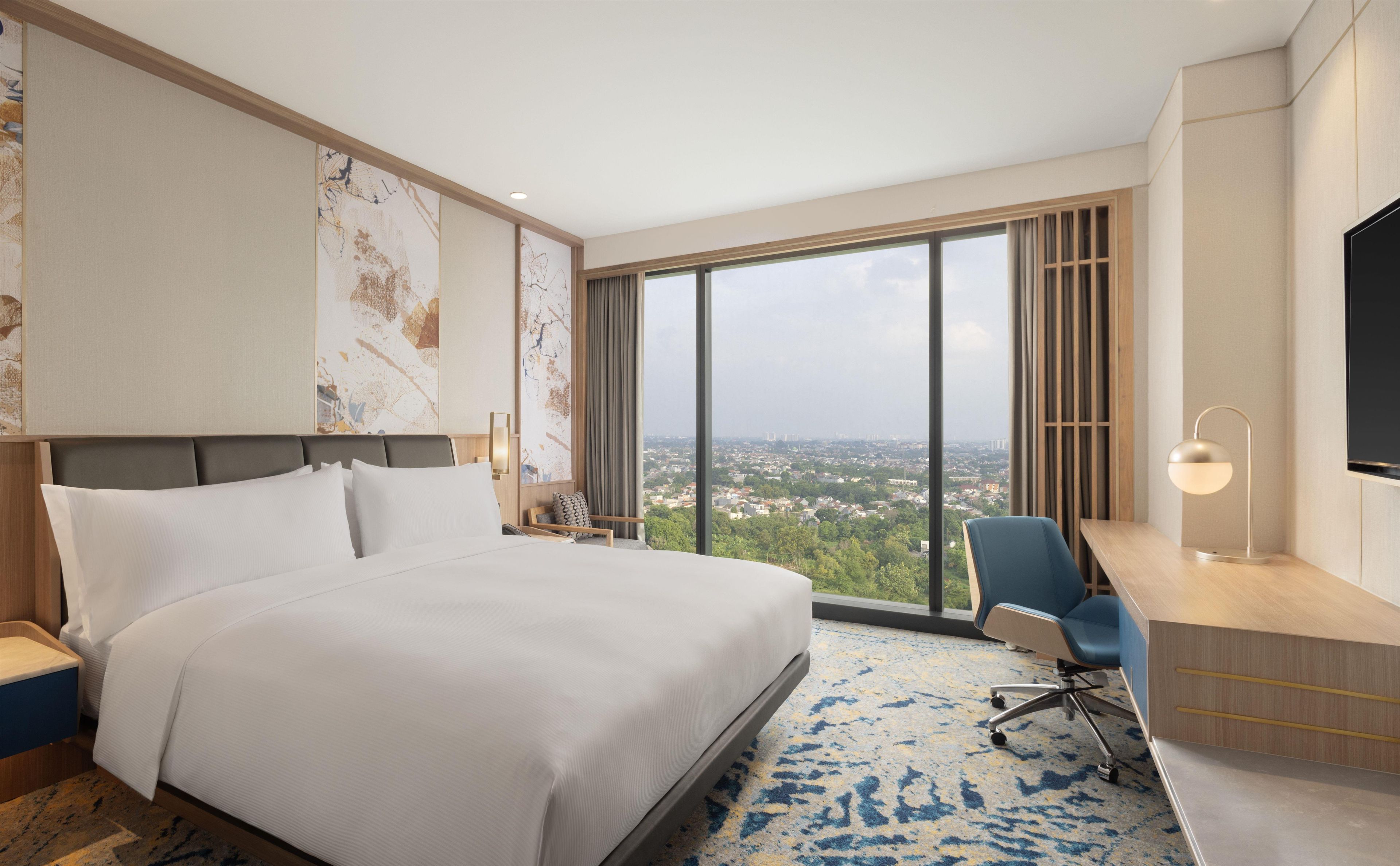 Habitación de hotel con una cama grande, un escritorio con silla, televisor y vistas a un paisaje urbano a través de amplias ventanas.