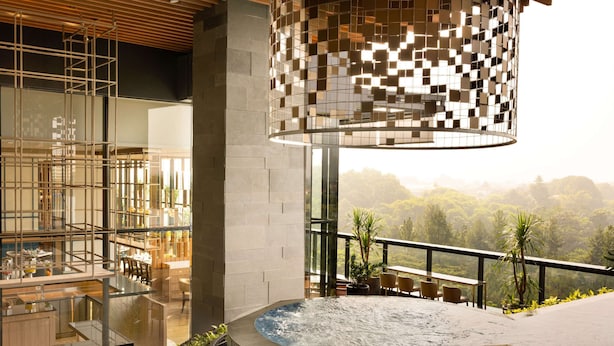 Un espacio moderno al aire libre con jacuzzi y vistas a un paisaje exuberante, rodeado por un cerramiento de vidrio y amueblado con taburetes y sillas.
