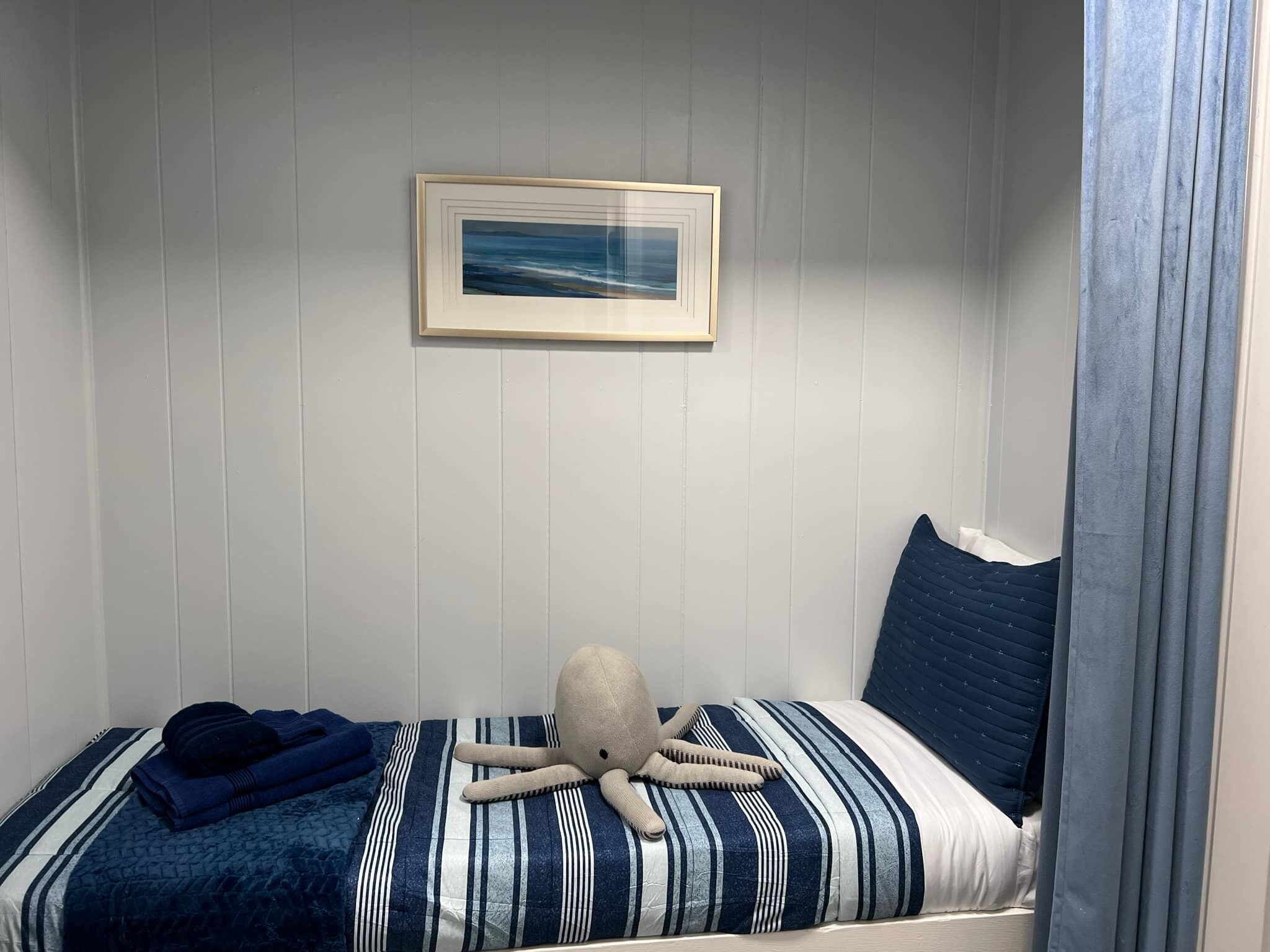 A small, single-bed room with a striped bedspread, a framed picture on the wall, and a curtain partially drawn to one side.