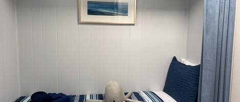 A small, single-bed room with a striped bedspread, a framed picture on the wall, and a curtain partially drawn to one side.