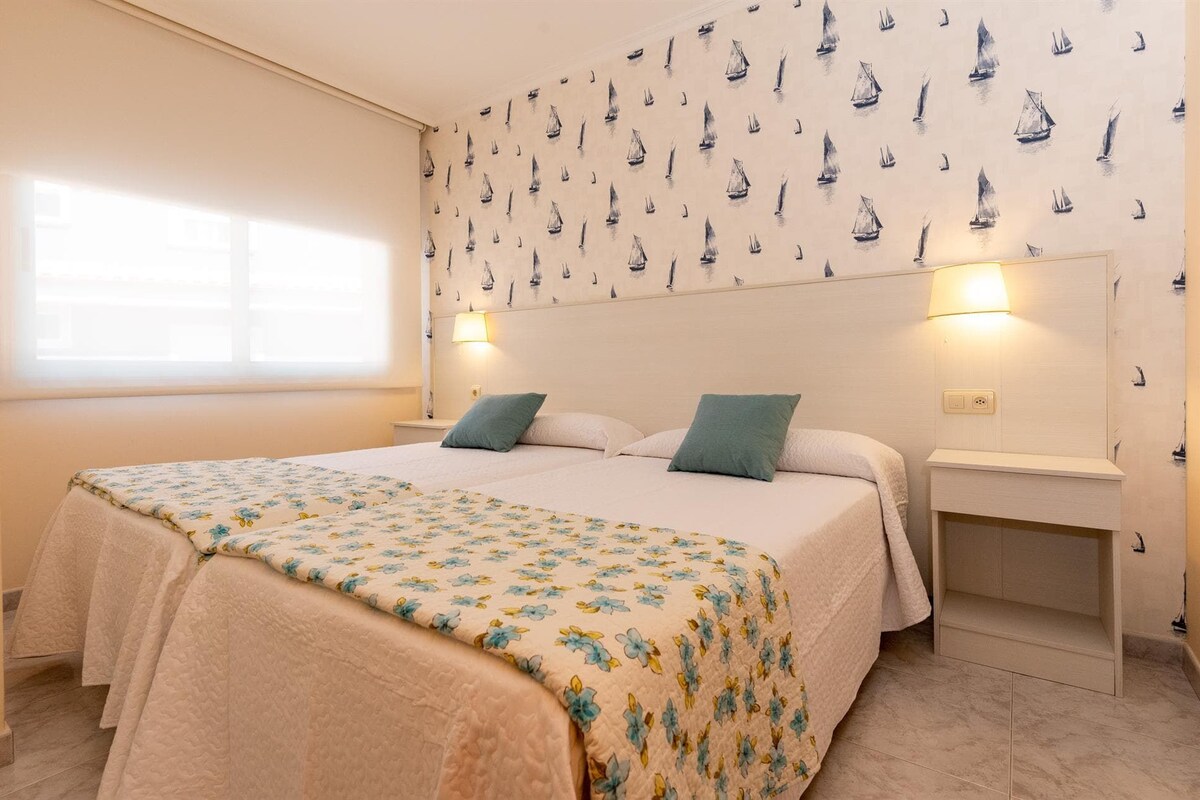 Slaapkamer met een bed, twee kussens, een nachtkastje, behang met zeilboten en een raam met jaloezieën.