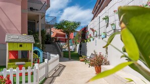 Un sendero estrecho bordeado por una valla de piquetes blancos, un parque infantil y un edificio con balcones y plantas en macetas.