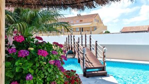 Una terraza de madera que conduce a una piscina con techo de paja y abundante vegetación.