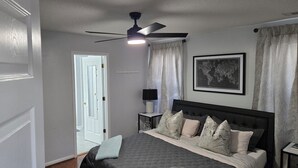 Un dormitorio con una cama, ventilador de techo, mesita de noche, lámpara, un cuadro enmarco y una puerta.
