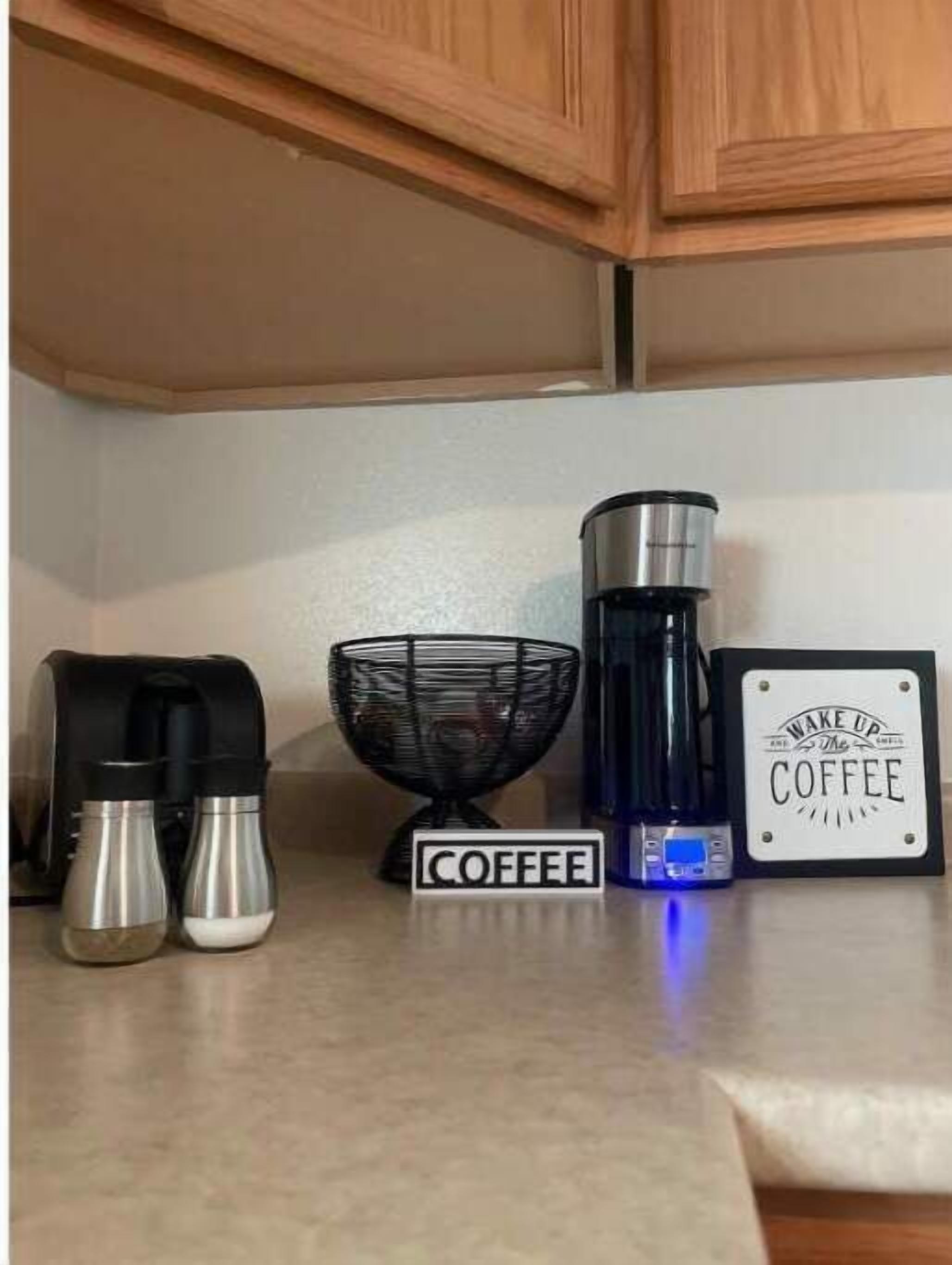 A kitchen counter with a coffee maker, two thermos flasks, a coffee grinder, and a sign that reads "Wake Up The Coffee".