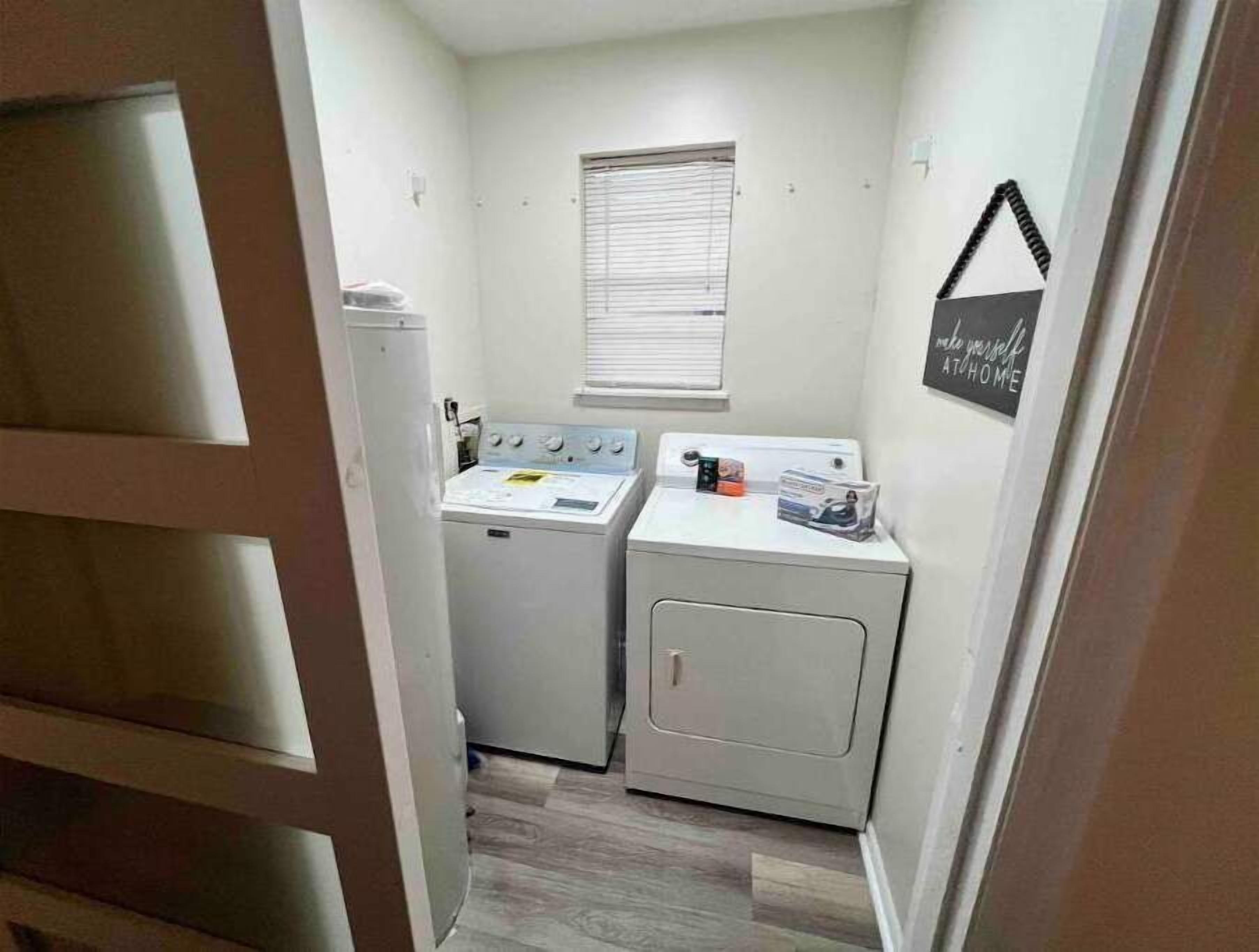 Un lavadero pequeño con dos lavadoras y una secadora, una ventana con persianas y un letrero motivacional en la pared.