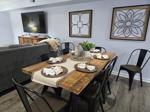A dining area with a wooden table set for four, surrounded by black chairs. There are framed artworks on the wall, a television above the sofa, and a plant on the table.
