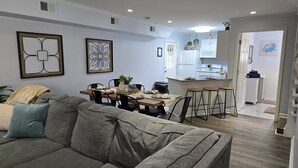 Un salon moderne avec un canapé gris, un coin repas avec une table en bois et une cuisine dotée de meubles blancs et d’un réfrigérateur en acier inoxydable.