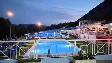 Et udendørs swimmingpool med et overdækket område, omgivet af et hvidt hegn og med et bjergrigt landskab i baggrunden.
