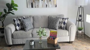 Un salon avec un canapé gris, des coussins à motifs, une table basse, une œuvre d’art murale, une plante en pot et une lampe sur pied.