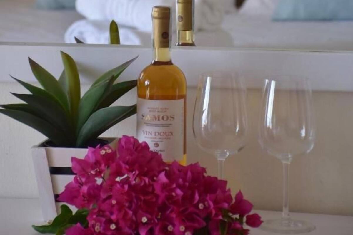 A bed with white bedding and light blue pillows, a bottle of wine, two empty glasses, and a potted plant with pink flowers.