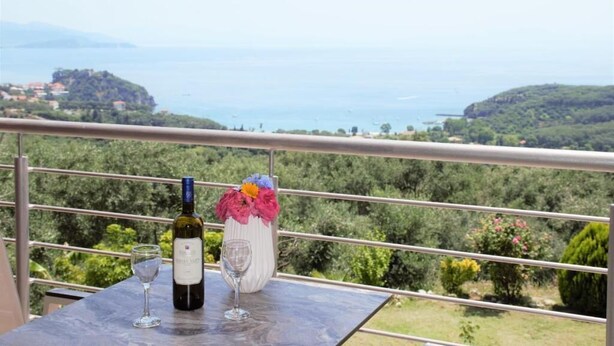 A balcony with a table, a bottle of wine, and a glass. The view includes a body of water and greenery. - Villa Thomas Nakas (Parga)