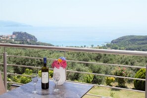 A balcony with a table, a bottle of wine, and a glass. The view includes a body of water and greenery.