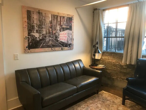 A living room with a black leather sofa, a large abstract painting, a lamp, and a view through a window with curtains.
