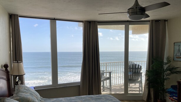 Ein Schlafzimmer mit großem Fenster zum Meer, einem Deckenventilator und einem Balkon mit Stühlen.