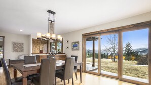 Un comedor amplio con una gran mesa de madera y sillas, una araña y vistas a un paisaje cubierto de césped a través de grandes puertas corredizas de vidrio.