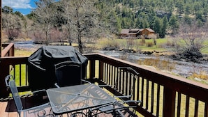 Una terraza con una parrilla, una mesa de metal y sillas, con vistas a un río y montañas boscosas.