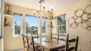 Un comedor con mesa y sillas de madera, ventanas amplias y una araña.