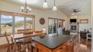 Una cocina amplia con una isla grande, una mesa de comedor de madera y sillas. Hay una chimenea de piedra, un ventilador de techo y ventanas grandes que ofrecen vistas al exterior.