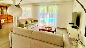 A modern living room with a sofa, coffee table, and a TV. There is a dining area with a table and chairs, and a kitchen in the background. - Villa Magellan — Charme, Espace & Plage à 250 m (Baie Orientale) (Orient Bay)