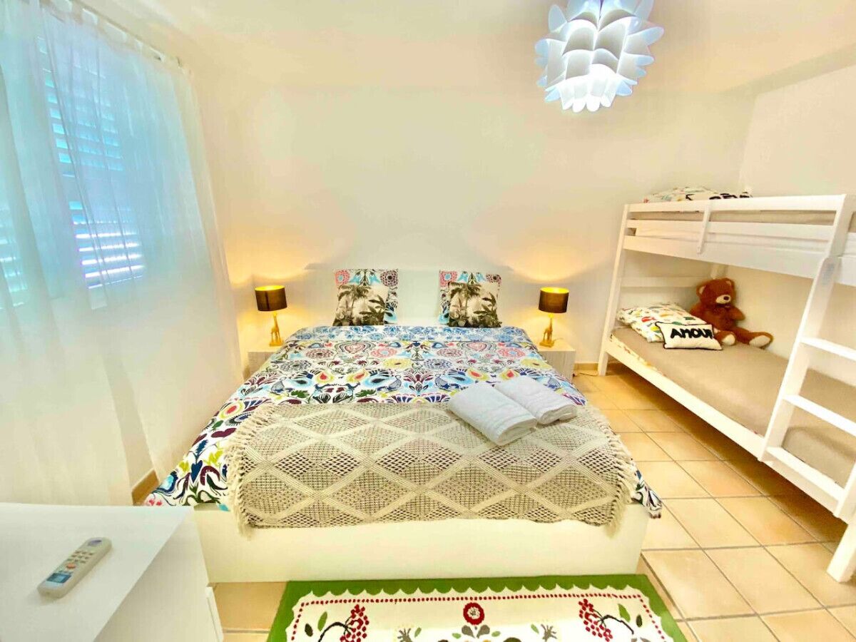 A bedroom with a bed, two bedside lamps, a bunk bed, a teddy bear, and a nightstand with a remote control.