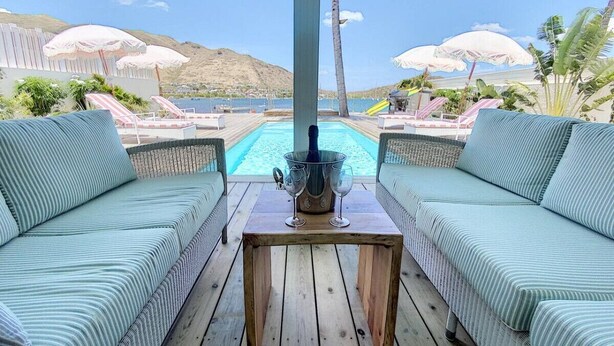 A wooden deck with two striped sofas facing a pool, a table with a drink, and umbrellas in the background. - Villa Horizon Lointain . Plage & piscine privée (Cul-de-Sac)
