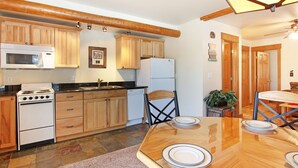 Une cuisine avec des meubles de rangement en bois, une cuisinière, un four à micro-ondes et un réfrigérateur. On y trouve une table à manger avec des assiettes et une chaise.