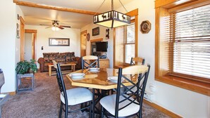 Un coin repas avec une table et des chaises en bois, un ventilateur de plafond et une grande fenêtre équipée de stores.