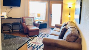 A living room with a brown leather sofa, a brown leather armchair, a small round table with a lamp, a ceiling fan, and a TV mounted on the wall. - Horsetooth Peak 26C - 1 Br Condo - Lake and Mountain Views (Estes Park)