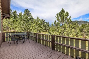 A wooden deck with a table and chairs, surrounded by a railing, overlooking a forested area with mountains in the background.