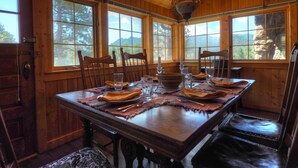 Comedor de madera con una gran mesa dispuesta para cuatro personas, flanqueada por ventanales altos que dan al exterior.