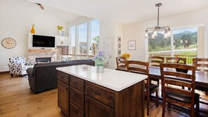 A modern kitchen with a central island, wooden dining table, and chairs. There is a TV on the wall and a large window providing a view of the outdoors.