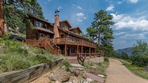 A large log cabin with multiple levels, a stone chimney, and a wooden deck. - Bishop's Retreat - Historic Log Home - Panoramic Mountain Views - 20-NCD0237 (Estes Park)