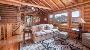 A log cabin interior with a wooden ceiling, a sofa, a patterned rug, and a view of mountains through large windows. - Bishop's Retreat - Historic Log Home - Panoramic Mountain Views - 20-NCD0237 (Estes Park)