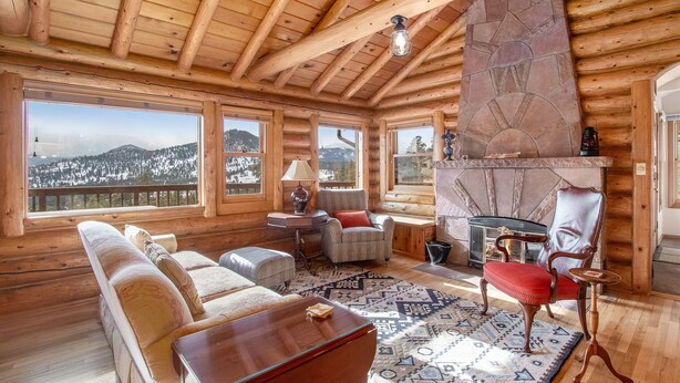 A log cabin interior with a stone fireplace, large windows, and a cozy seating area. - Bishop's Retreat - Historic Log Home - Panoramic Mountain Views - 20-NCD0237 (Estes Park)
