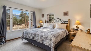 A bedroom with a large bed, two bedside tables with lamps, a desk, a chair, and a window with a view of trees and mountains.