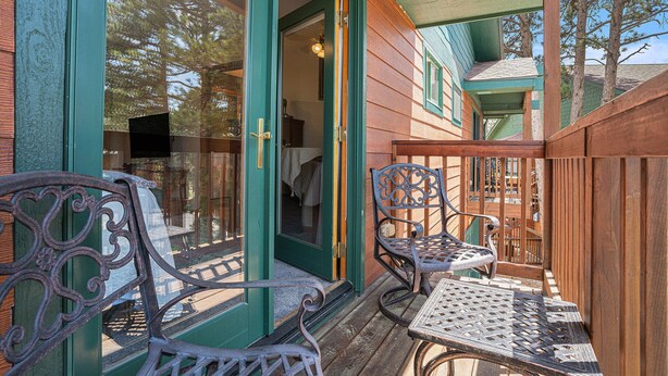 Une terrasse en bois avec deux chaises en métal et une table, une porte verte, et une vue sur des arbres et un bâtiment en arrière-plan.
