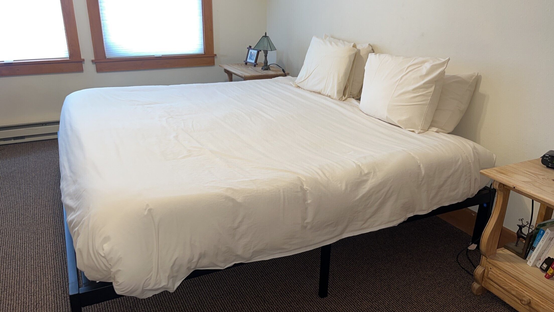 Una cama bien tendida con sábanas blancas en una habitación con ventana, una mesita con lámpara y una estantería.